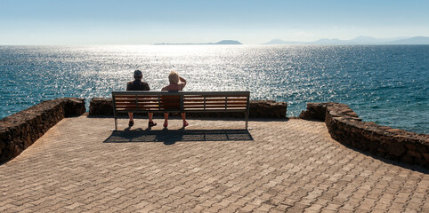 A senior couple sitting on a bench on the beach in the sun looking out to sea.