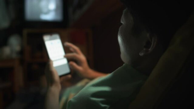 Over-shoulder shot of Chinese man or woman lying on bed in tiny micro flat, holding smartphone and browsing on internet, while watching movie on TV and relaxing at home after workday
