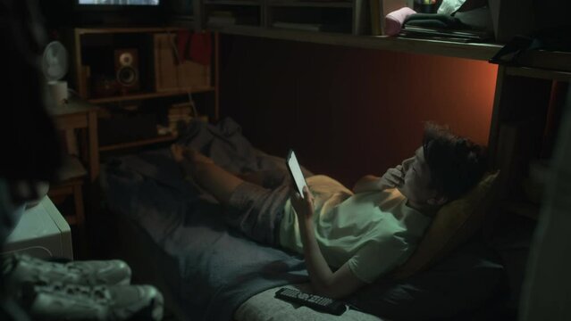 Full shot of tired young Chinese androgynous woman lying on bunk bed in cramped tiny rental flat, typing on smartphone and yawning, while resting at night after work, and movie playing on TV