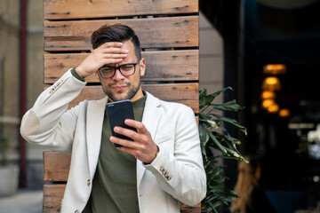 Overworked businessman in problems, feeling confused while looking at mobile phone outdoors.