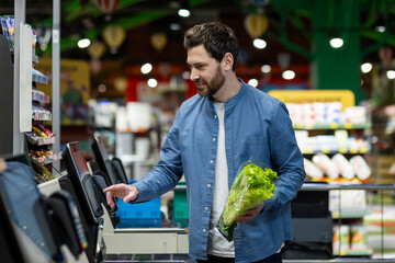 Adult male in a blue shirt using a self-checkout machine in a supermarket, holding fresh lettuce.