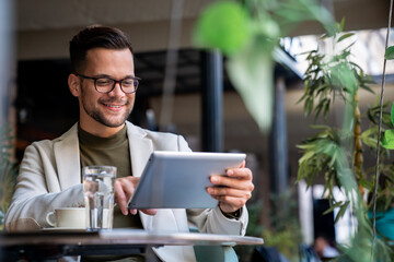 Stylish happy modern guy using tablet while sitting in coffee shop. Photo of a male working on a...