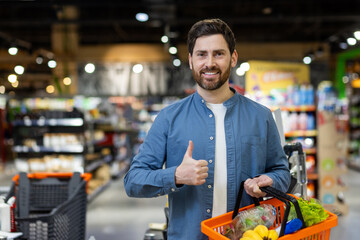 A man in a supermarket with a full shopping basket gives a thumbs up, signifying a positive...