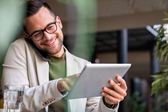 Smiling young business man sitting in Cafe sharing business ideas with colleague over mobile phone while using digital tablet.
