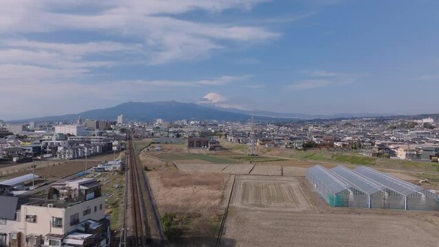 Forwards fly above train line and buildings in town. Large greenhouses on bank of river. Mountains with iconic Mount Fuji peak in distance. Japan