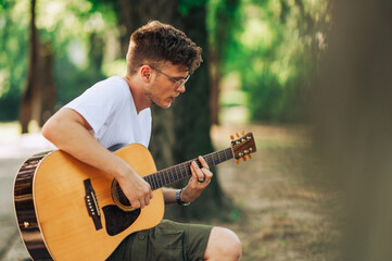 A young man is sitting in city park and playing a guitar.