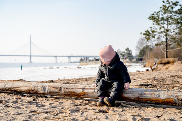 Toddler child sits on a log on a sandy beach in St. Petersburg during the cold season