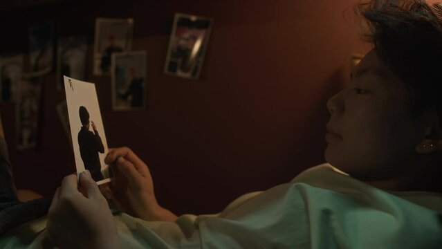 Medium close-up shot of young Chinese girl lying on bunk bed in tiny capsule flat at nighttime and looking longingly at photo of her boyfriend