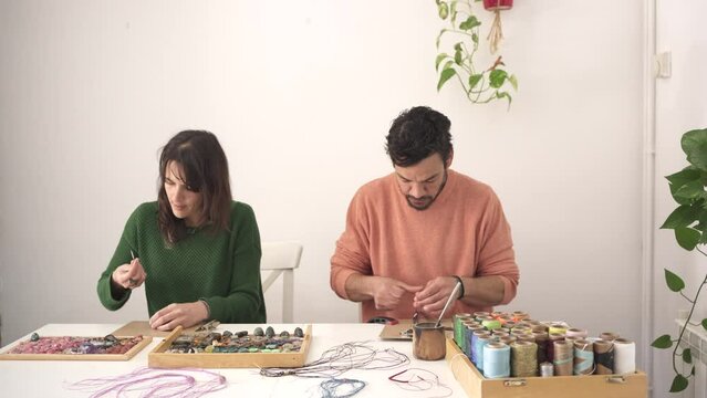 Couple making crafts with stones and macram&eacute;. Woman in green sweater filing gem and man weaving bracelet in salmon orange sweater.