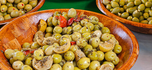 A close up of Mediterranean oil dressed green olives in a wooden bowl with herbs, sun-dried tomatoes and lemon wedges for sale at a market..