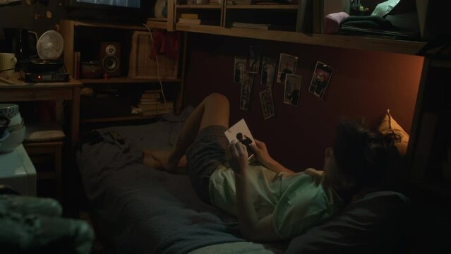 Full shot of young Asian woman or man lying on bunk bed in dark cluttered capsule flat at nighttime, watching movie on TV, then taking photo of boyfriend from wall and looking at it longingly