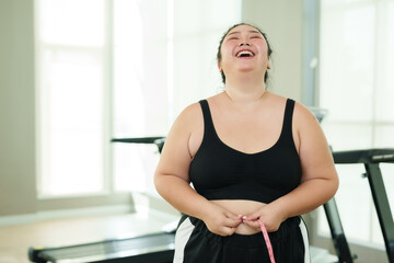 Chubby Asian woman exercise in gym, Happy smile in joy progress with tape measure, triumph in health and fitness journey. Elated female measuring waist, a victorious moment in her workout regimen