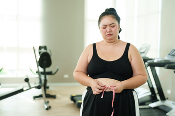 Chubby Asian woman exercise in gym, Pensive measuring waistline, reflecting on fitness journey, Contemplative female with tape measure, considering health progress, in thoughtful gym setting