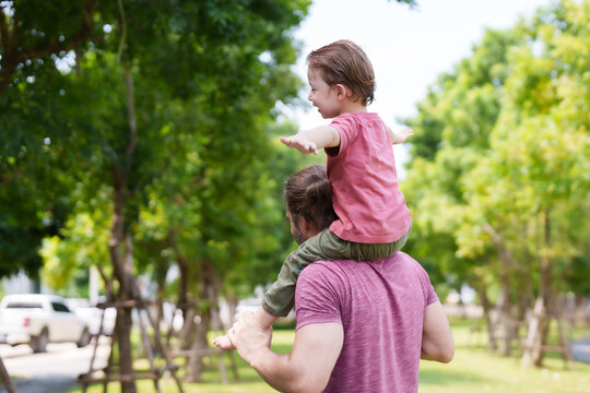 Male with beard gives young child piggyback ride in lush park, arms outstretched, embodying freedom and child’s glee amidst verdant trees. Bearded elder supporting small youngster on back,.