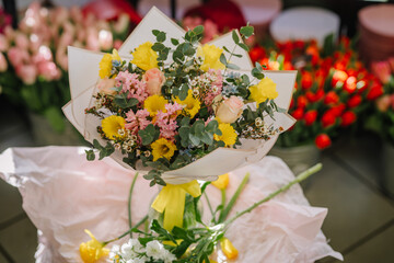Valmiera, Latvia - March 7, 2024 - A bouquet of yellow, pink, and cream flowers wrapped in paper sits on a table with red tulips in the background.