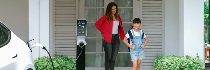 Happy little young girl learn about eco-friendly and energy sustainability as she help her mother recharge electric vehicle from home EV charging station. EV car and modern family. Panorama Synchronos