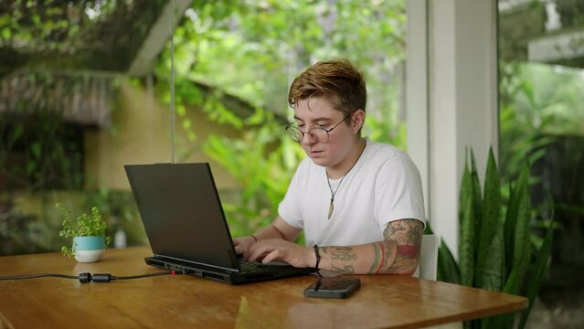 Transgender pro types on laptop in bright, plant-filled office. Engrossed in work, tattooed individual enjoys peaceful, productive environment. Inclusion, tech industry diversity represented. Slowmo