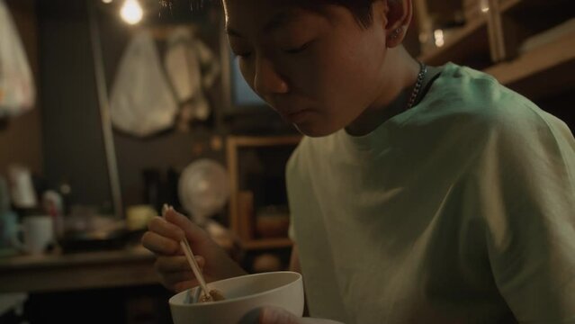Medium close-up shot of hungry young Chinese woman with short hair eating stir-fry out of bowl with chopsticks and liking it, while resting in her micro apartment in evening after work