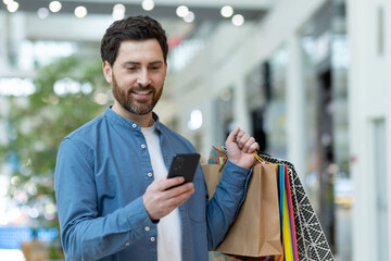 A modern shopper using his phone while carrying multiple shopping bags in a busy mall setting,...