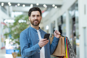 Smiling male shopper using a smartphone while holding colorful shopping bags in a bright shopping mall setting.