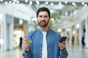 Happy, bearded man holding a credit card and smartphone with a content smile, standing in a shopping mall with blurred background.