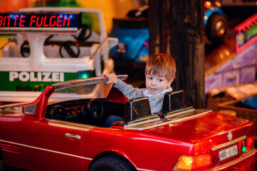 A cheerful toddler beams as he takes the wheel of a bright red convertible on a merry-go-round,...