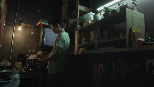 Full shot of young Chinese man or woman standing in dark shabby tiny apartment, stirring food cooking in steaming pot on small electric stove, and watching movie on TV, while making dinner after work