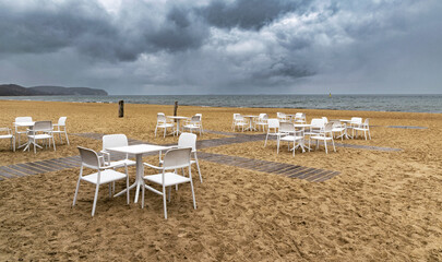 Fototapeta na wymiar White lounge chairs and table on the sandy beach with stormy sea