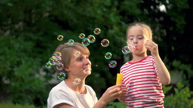 Little girl and mother blow soap bubbles together in green city park. Mother with joyful little girl plays soap bubbles walking in park. Little girl and mother rest blowing soap bubbles in garden