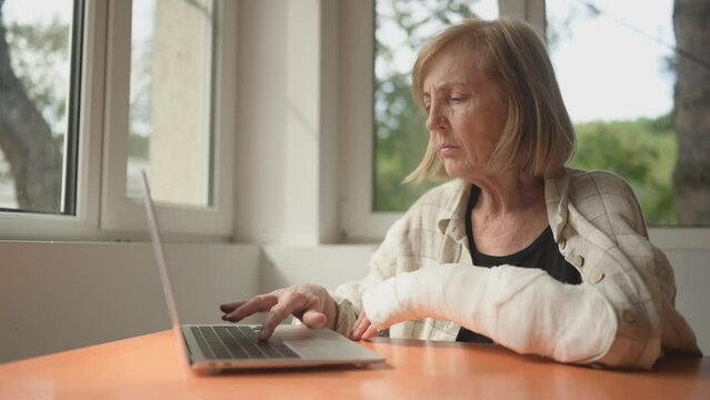 Senior elderly woman working at home with laptop computer with injury broken arm in cast indoors in front of windows. Old lady touching wrist in gypsum sling bandage feeling hurt. 