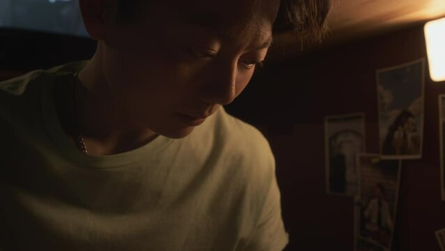 Close-up shot of young androgynous Chinese female student with short hair sitting in bedroom at night, reading textbook, revising and learning material for university exam, movie on TV in background