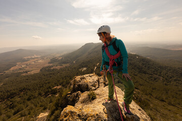 A woman is standing on a mountain top, wearing a helmet and a blue jacket. She is looking out over the landscape, possibly taking in the view or preparing for a climb