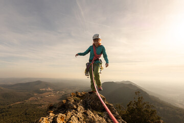 A woman is standing on a mountain top with a rope attached to her. She is wearing a helmet and a...