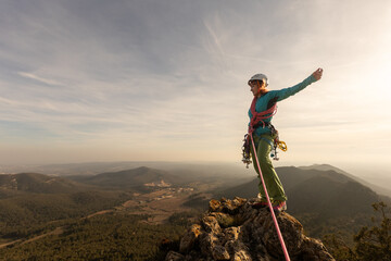 A woman is standing on a mountain top, wearing a blue jacket and a red harness. She is smiling and...