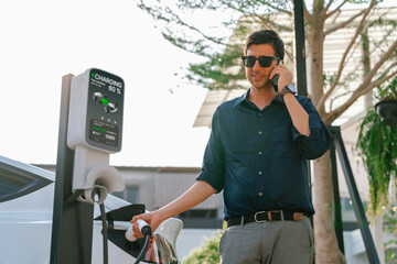 Young man recharge EV electric vehicle at green city park parking lot while talking on phone....