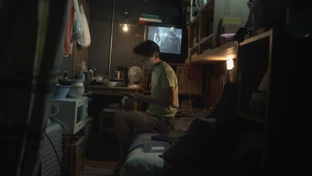 Full shot of young Chinese woman or man sitting on bunk bed in cluttered tiny flat with dim light, eating takeaway food with chopsticks, scrolling on smartphone, movie on TV in background