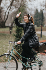 Young attractive woman wearing headphones enjoying a bike ride, waving hello with a smile in an urban park.