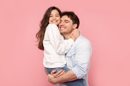 My best daddy. Happy daughter embracing her father as man holds girl in arms, both smiling, posing over pink studio backdrop
