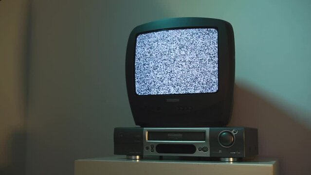 An old TV turns on a gray screen with black and white noise. An old TV with a video recorder stands on the bedside table in the living room and shows television noise. 