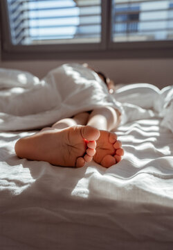 Feet of little sleeping child sticking out from under the blanket at sunny morning. Focus on feet