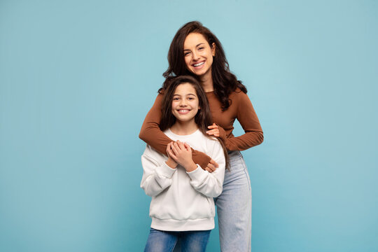 Mother's love concept. Happy young mom posing with her preteen daughter from the back, standing on blue studio background, both smiling at camera