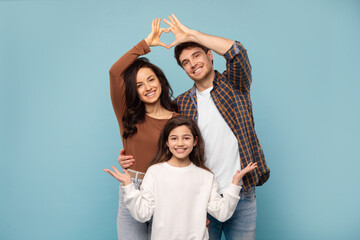 Happy parents joining arms making fingers heart above their daughter, posing on blue studio...