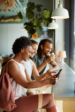 Three diverse friends sit at a high table by a window in a cafe after a yoga lesson. They drink coffee and look at mobile while laughing together