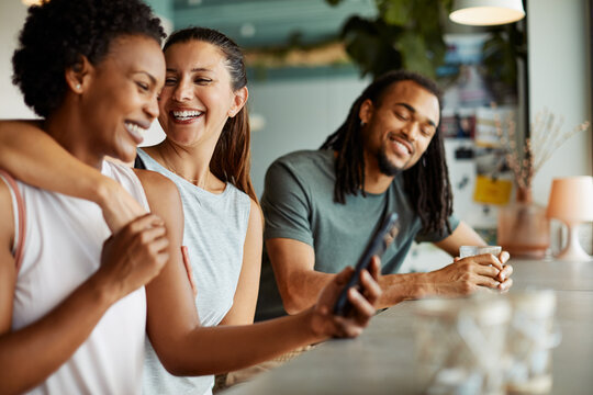 Three diverse friends laugh and fool around in a cafe after a yoga class. They are looking at funny pictures on a cell phone