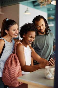 Three diverse friends sit together in a cafe in sportswear after a yoga class. They laugh together while looking at pictures on a mobile phone.