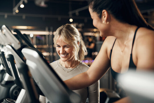 Female instructor laughing while helping a fit young woman riding on a stationary bike during a cardio exercise session in a gym