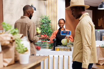 Black woman runs eco friendly store, offering organic groceries. She assists clients with a smile, providing a variety of fresh fruits and vegetables with sustainable packaging.