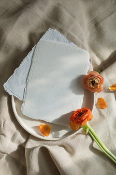 Overhead view of blank handmade paper cards on a ceramic plate with orange ranunculus flowers on beige tablecloth