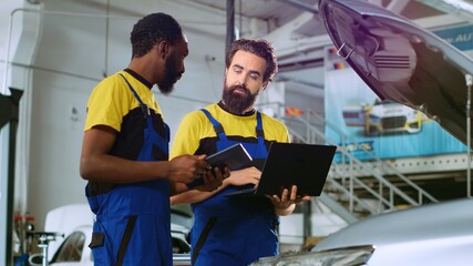 Mechanics in car service ordering new parts for damaged vehicle using laptop and tablet. Coworkers...