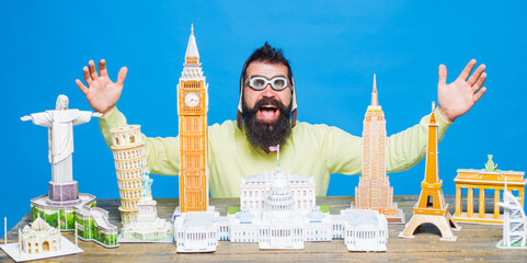 Travel, adventure, vacation, worlds monuments. Happy bearded man in pilot helmet and glasses near table with miniature copy of world architectural landmarks. Miniature buildings models. Man traveling.